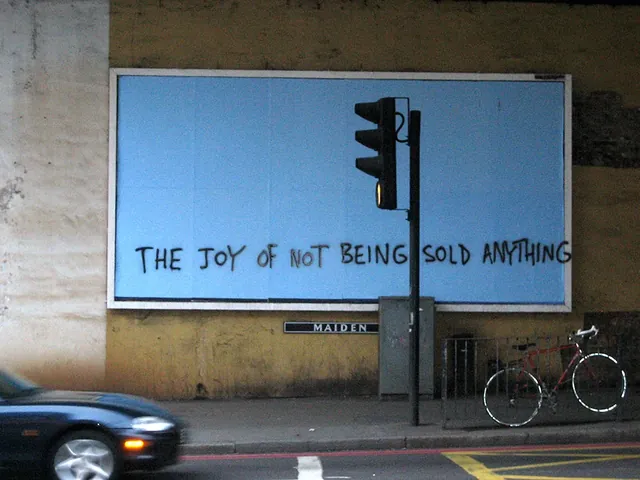 A blank blue billboard under a London railway bridge with THE JOY OF NOT BEING SOLD ANYTHING painted across it